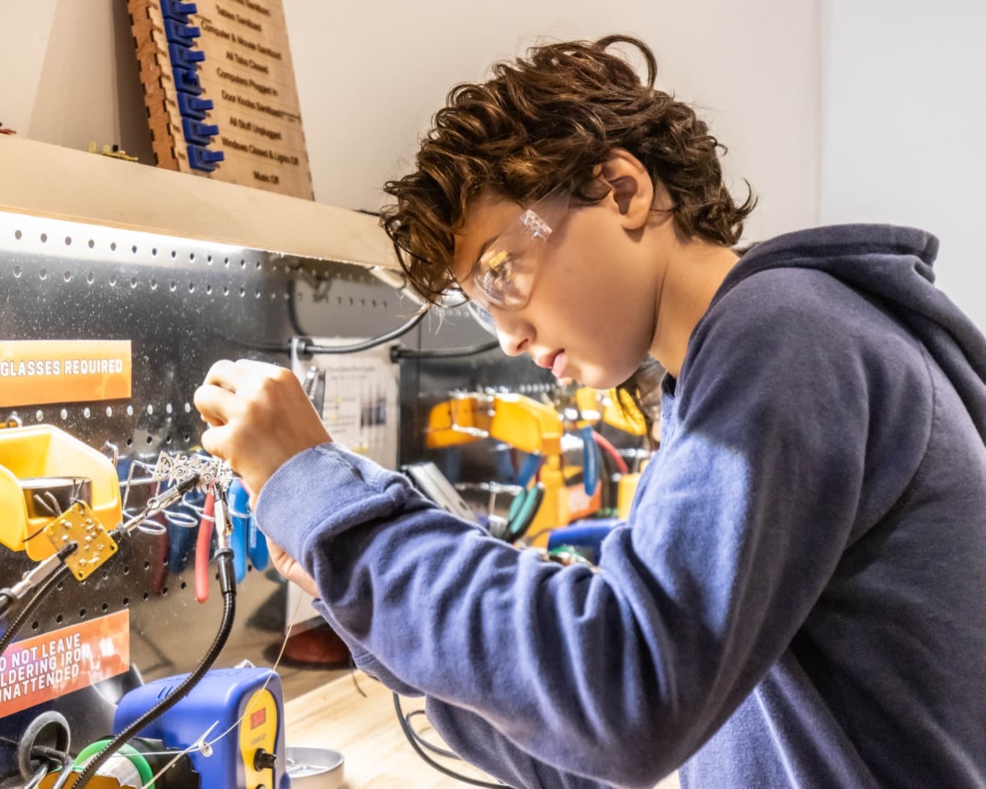 A kid in safety glasses soldering electronics at a real workbench.